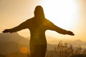A woman stands with arms open, embracing the sunrise in a mountainous landscape, creating a tranquil silhouette.